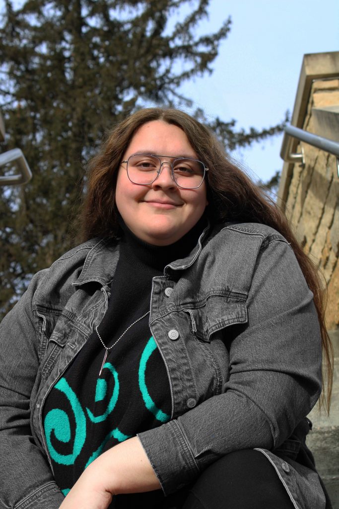 A woman with long hair wearing glasses and a black denim jacket sitting on steps with tress and a blue sky in the background
